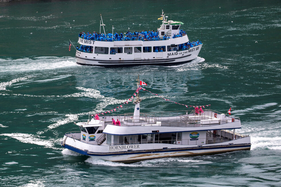 Американската туристическа лодка Maid Of The Mist с ограничение до 50% заетост по правилата на щата Ню Йорк плава покрай канадско корабче, което работи по правилата на Онтарио само до шест пътници на борда в Ниагарския водопад, Онтарио, Канада.
