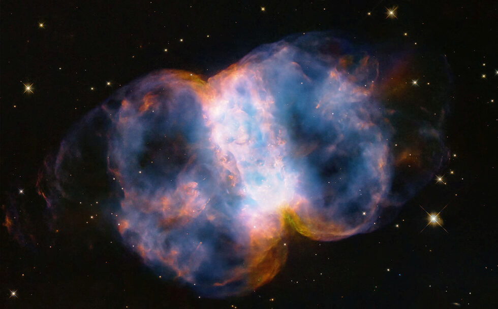 Мъглявината "Малкият дъмбел", известна още като Messier 76 или M76, разположена на 3400 светлинни години в северното кръгово съзвездие Персей, се вижда на изображение, направено от телескопа "Хъбъл" на 23 април 2024 г.<br /><br />Името "Малкият дъмбел" идва от формата на двулицевата му структура от цветни, пъстри, светещи газове, наподобяваща балон, който е притиснат около средната част на талията. Подобно на надуващ се балон, лобовете се разширяват в пространството от умираща звезда, която се вижда като бяла точка в центъра.<br /><br />Смазващата ултравиолетова радиация от свръхгорещата звезда кара газовете да светят. Червеният цвят е от азота, а синият - от кислорода. Вижда се и умираща звезда, като бяла точка в центъра.